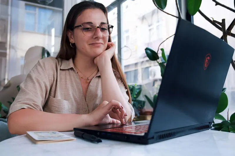 Mujer con gafas apoyando la barbilla en la mano mientras reflexiona frente a su portátil y una libreta